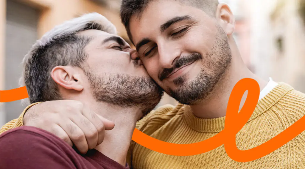 Two men share a tender kiss and hug on a city street, wearing sweaters.