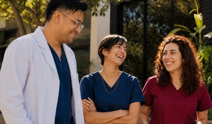 Four healthcare workers stand outside a wellness center, smiling and chatting in scrubs and a white coat.
