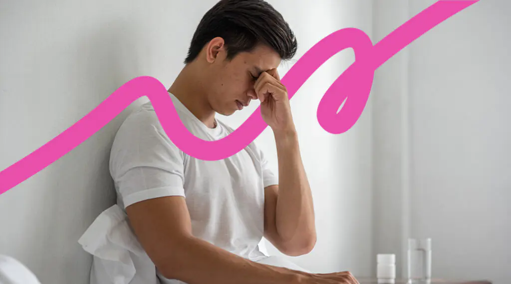 Young man in a white t-shirt sits against a wall, head in his hand, looking stressed or fatigued.