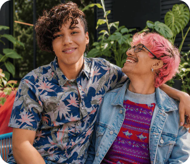 Two friends sitting close together outdoors, smiling at the camera; one wears a tropical print shirt and the other a denim jacket with pink hair.
