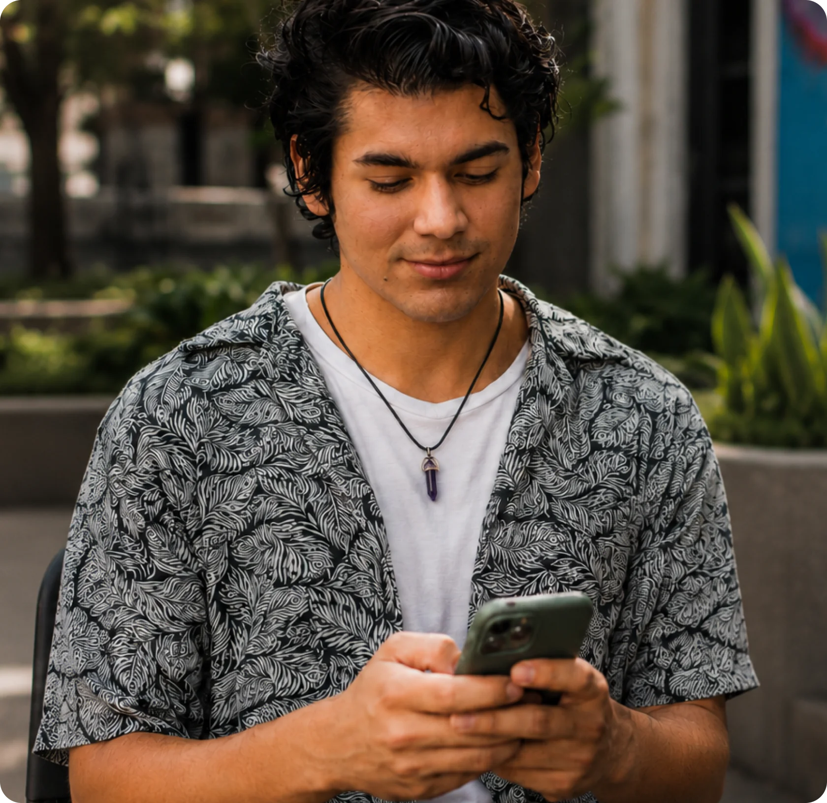 Man outdoors, looking down at his smartphone while seated, wearing a patterned short-sleeve shirt and necklace.